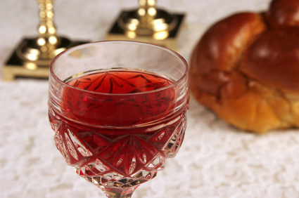 A Shabbat table with a glass of wine, gold candlestick holders and challah in the background.